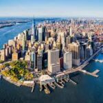 Aerial view of Manhattan's waterfront with dense skyscrapers and blue harbor water in the foreground, showing One World Trade Center in the distance.